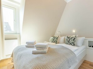 A bedroom with a bed, pillows, a bedside table, and a window. - Holiday Home Heide (Sylt)