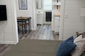 A room with a bed, a TV mounted on the wall, a dining area with a table and chairs, and a kitchen area with cabinets and a window. - Atlanta South Cabin Getaway, a Cozy Stay Near I-75 (McDonough)