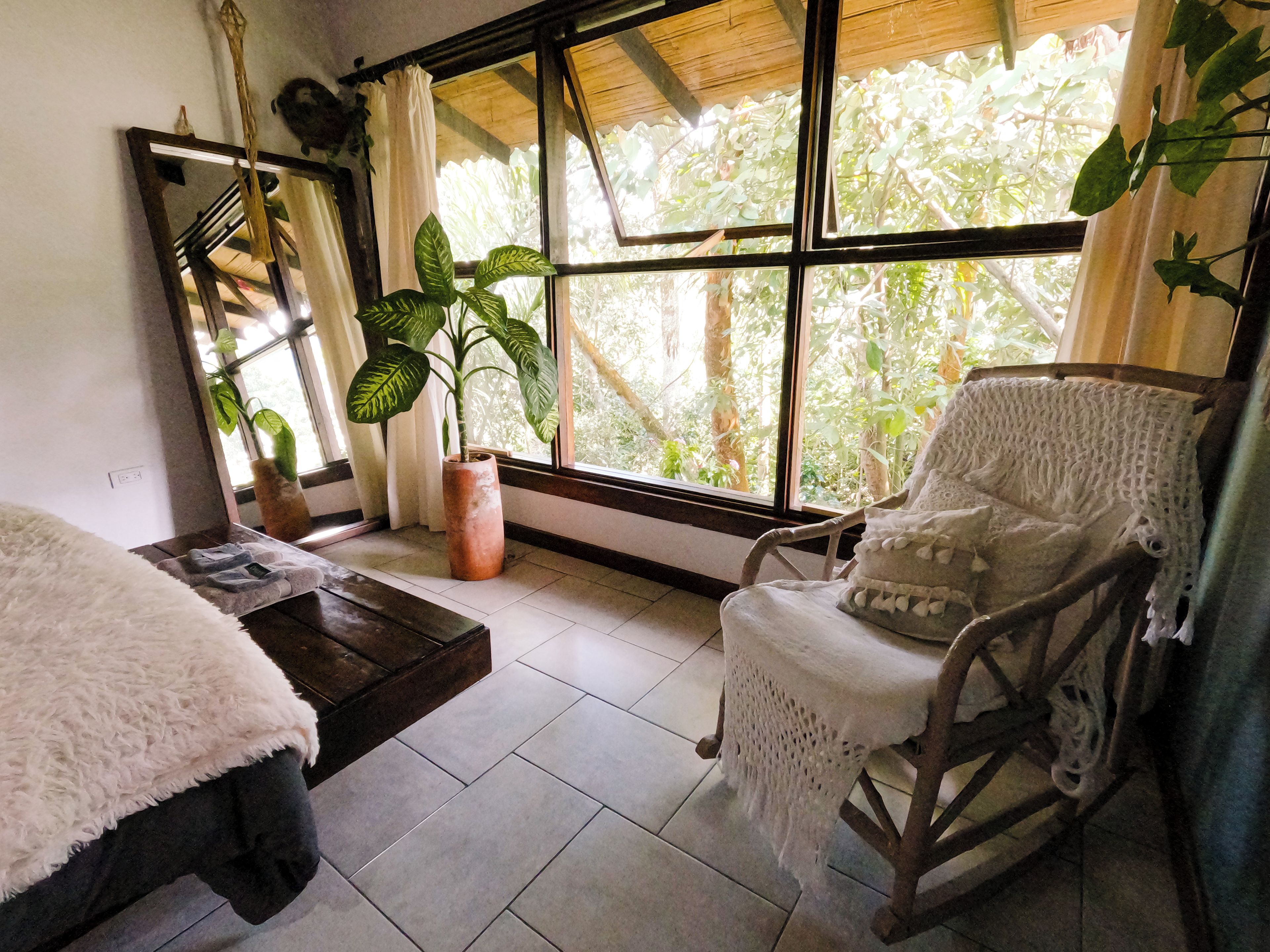 Una habitación con un ventanal amplio, una mecedora, una mesita pequeña y una planta en maceta.