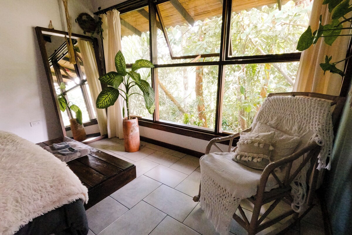 Una habitación con un ventanal amplio, una mecedora, una mesita pequeña y una planta en maceta.