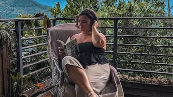 Una persona sentada en una silla de mimbre en un balcón con vistas a montañas y vegetación.