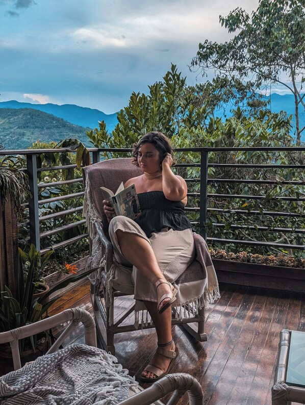 A person seated on a wicker chair on a balcony with a view of mountains and greenery.