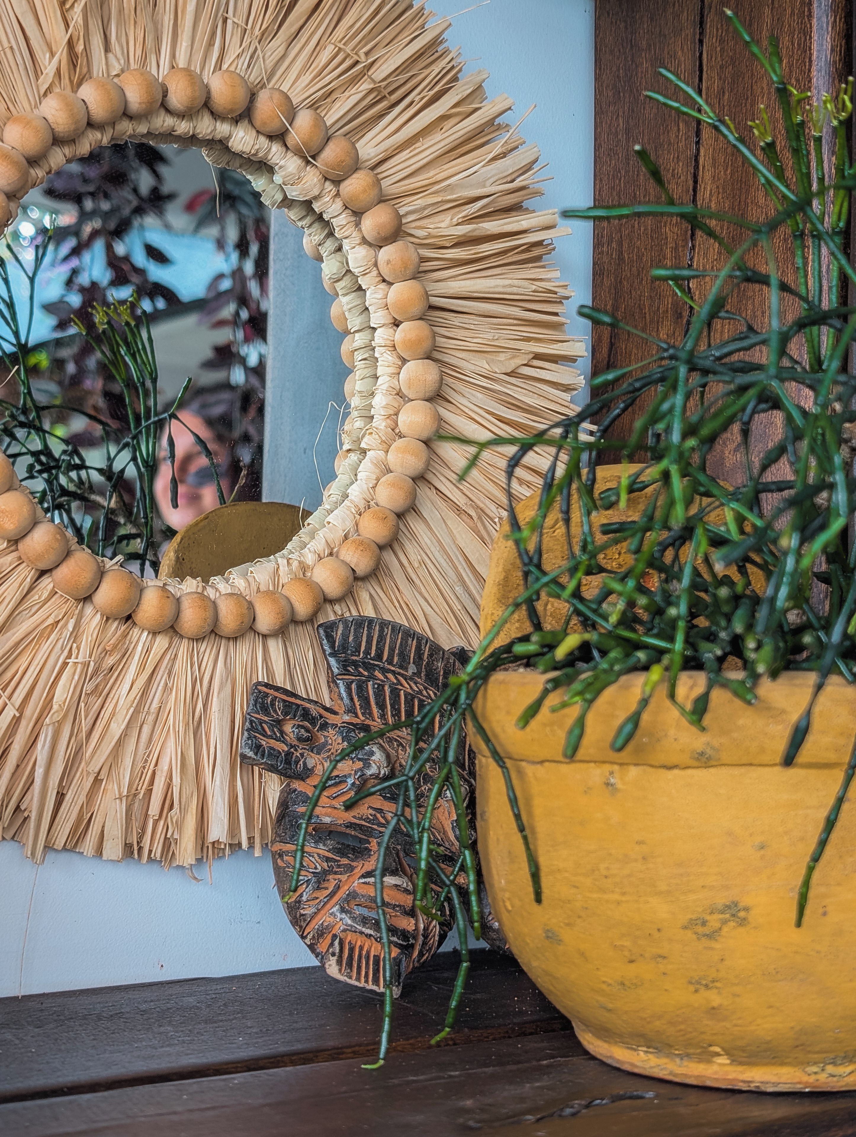 Un espejo decorativo con un borde de paja y cuentas, una pequeña figura tallada en madera y una planta en maceta con follaje verde.
