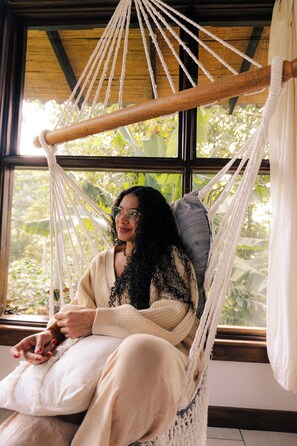 A hammock chair with a wooden frame and white ropes, positioned in front of a window with a view of greenery.