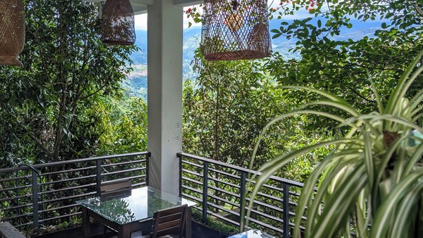 Un área de comedor al aire libre con una mesa y sillas de vidrio, rodeada por una barandilla, con vista a follaje y montañas al fondo.