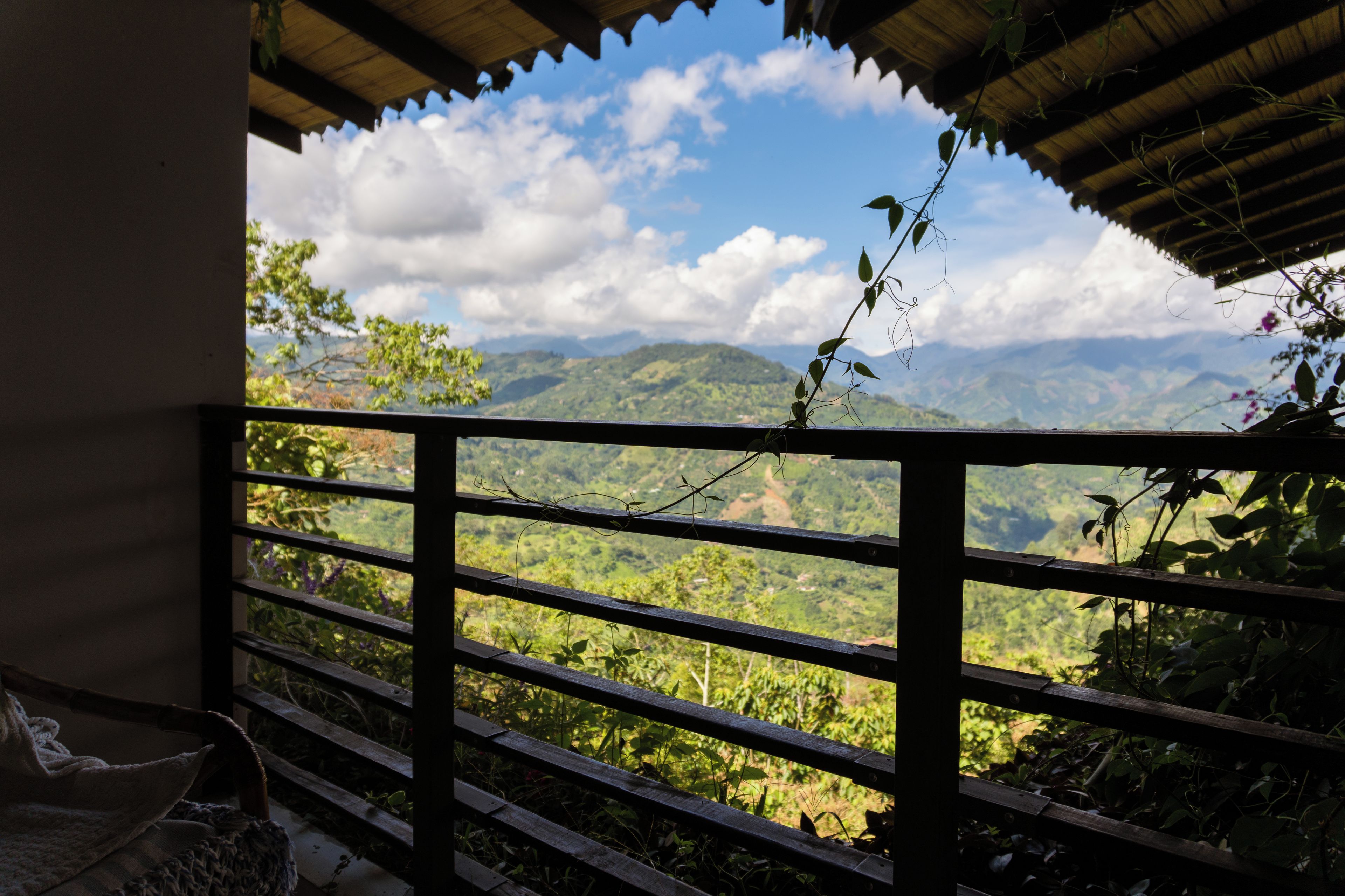 Vista desde un balcón con baranda de madera, sobre un paisaje montañoso con vegetación y cielo despejado.