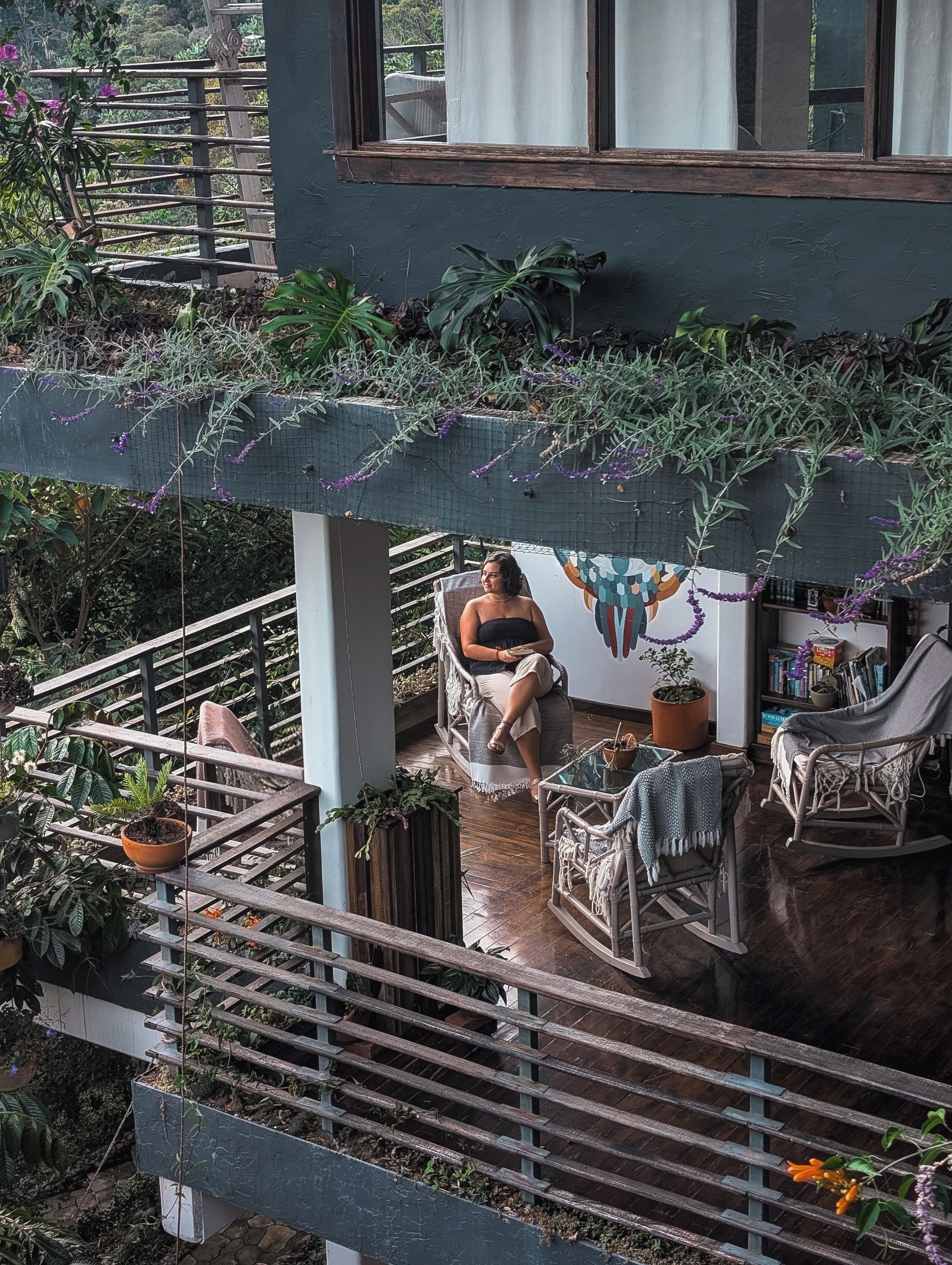 A balcony with a person seated on a chair, surrounded by potted plants and a wooden railing.