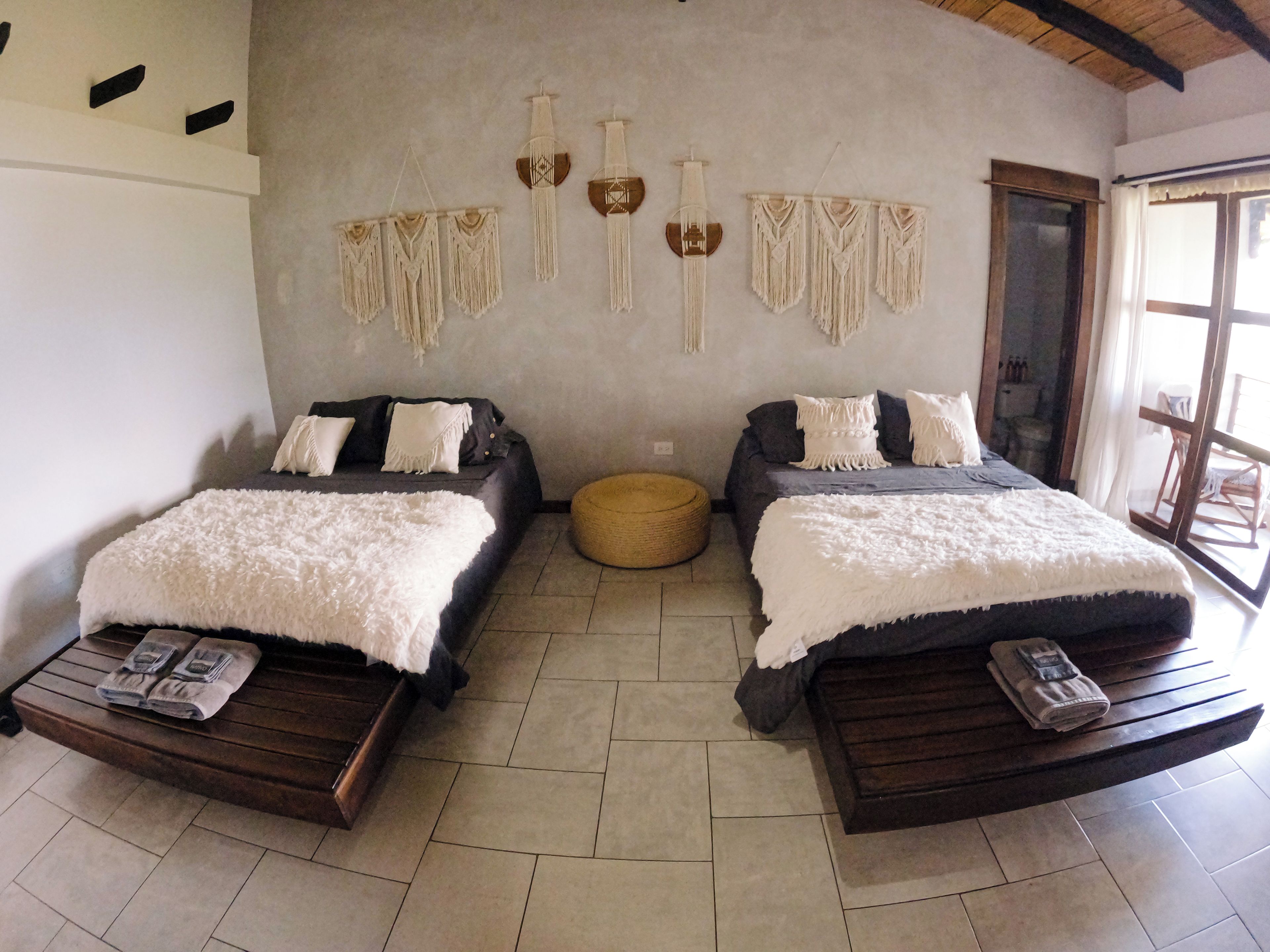 A room with two beds, a round ottoman, and macramé wall hangings.