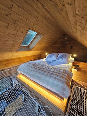 A cozy wooden cabin bedroom with a sloped ceiling, a skylight, a bed with a white blanket, and a small bedside table.