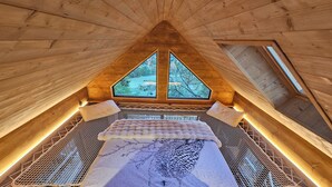 Habitación de madera con techos altos, una cama, dos ventanas y vista a árboles.