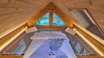 A high-ceilinged wooden room with a bed, two windows, and a view of trees.