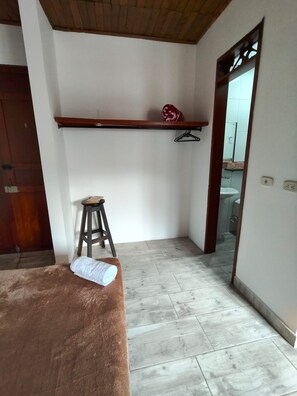 A room with a wooden ceiling, a brown sofa, a small stool, a wooden shelf with a red object, and a bathroom visible through an open door.