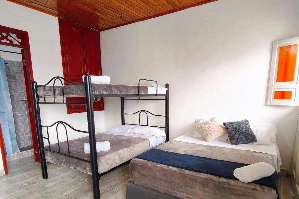 A room with a bunk bed, a single bed, wooden flooring, a red door, and a hanging light fixture.