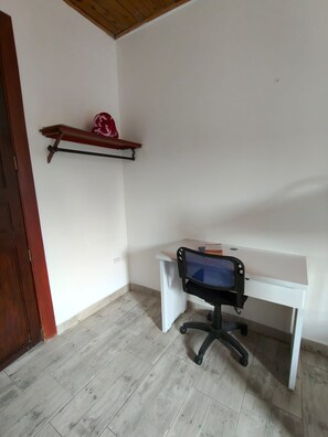 Una habitación con un escritorio blanco, una silla de oficina negra, una estantería de madera con un objeto decorativo y una puerta de madera.