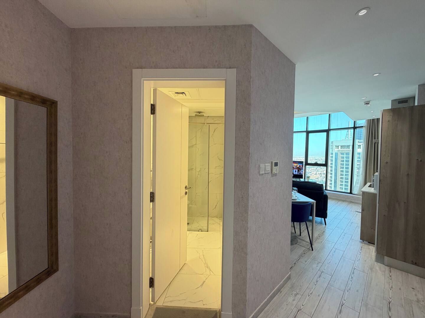 Un interno moderno con un corridoio che conduce a un bagno con porta in vetro, un'area pranzo con tavolo e sedie, e ampie finestre con vista sulla città.