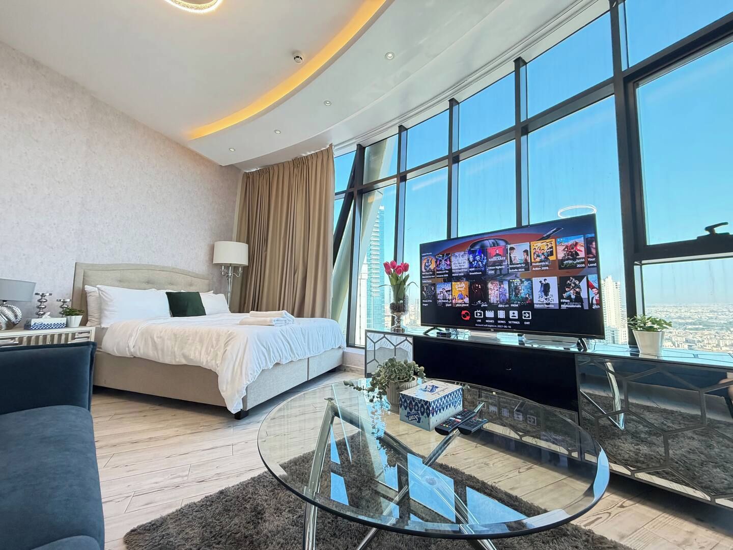 A modern hotel room with a large bed, a glass coffee table, a flat-screen TV, and a city view.