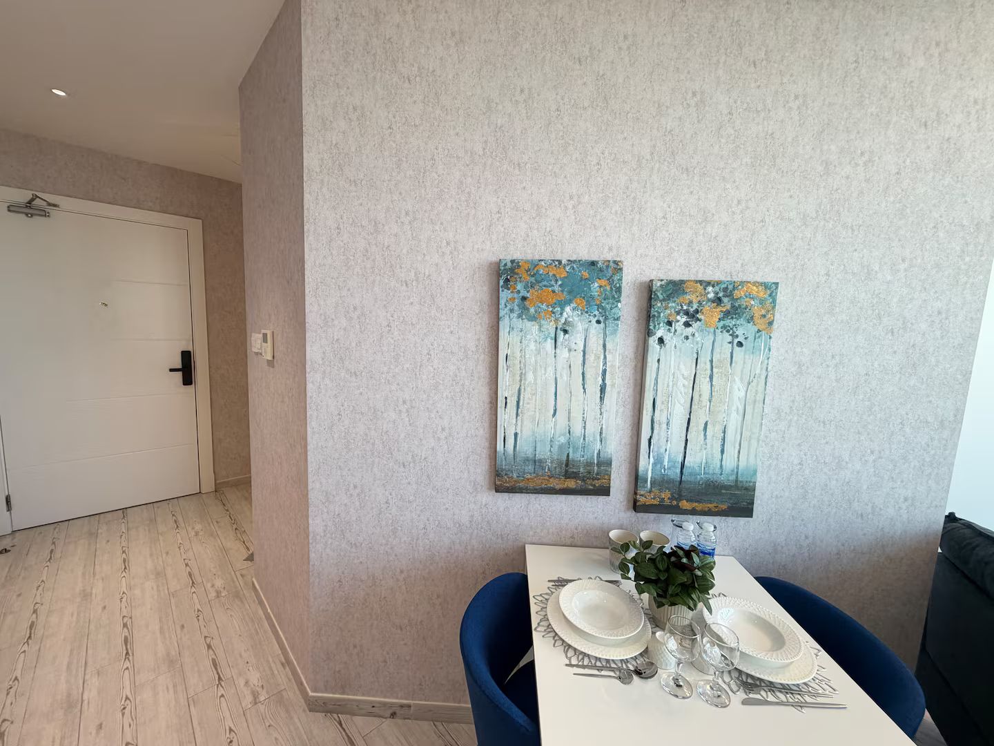 A dining area with a white table set for two, two paintings on the wall, and a door leading to another room.