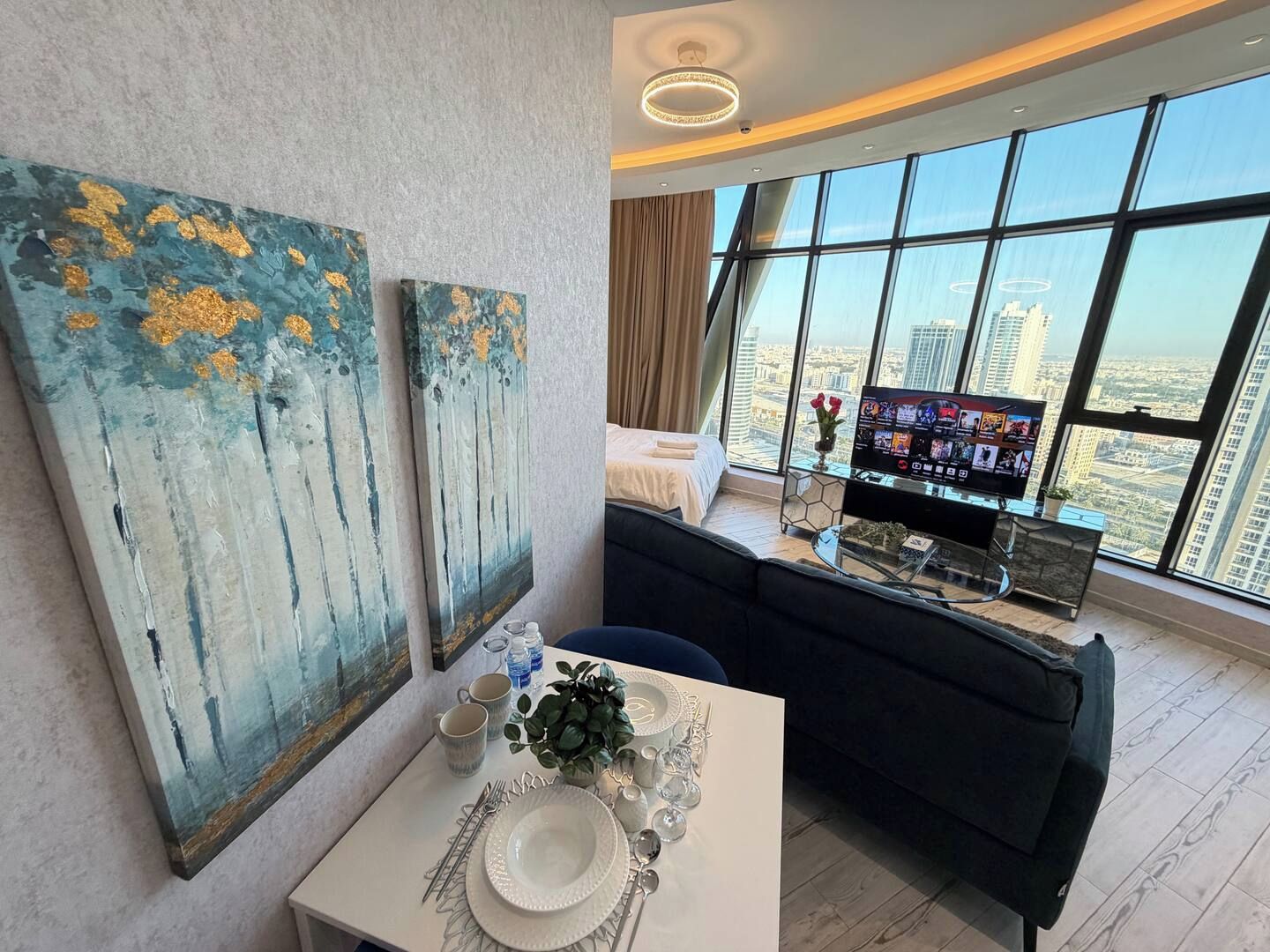 A modern hotel room with a dining area, a sofa, a TV, and large windows offering a city view.