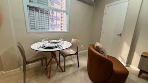 Uma área de refeições com uma mesa redonda posta para dois, uma janela com persianas e uma porta à direita.