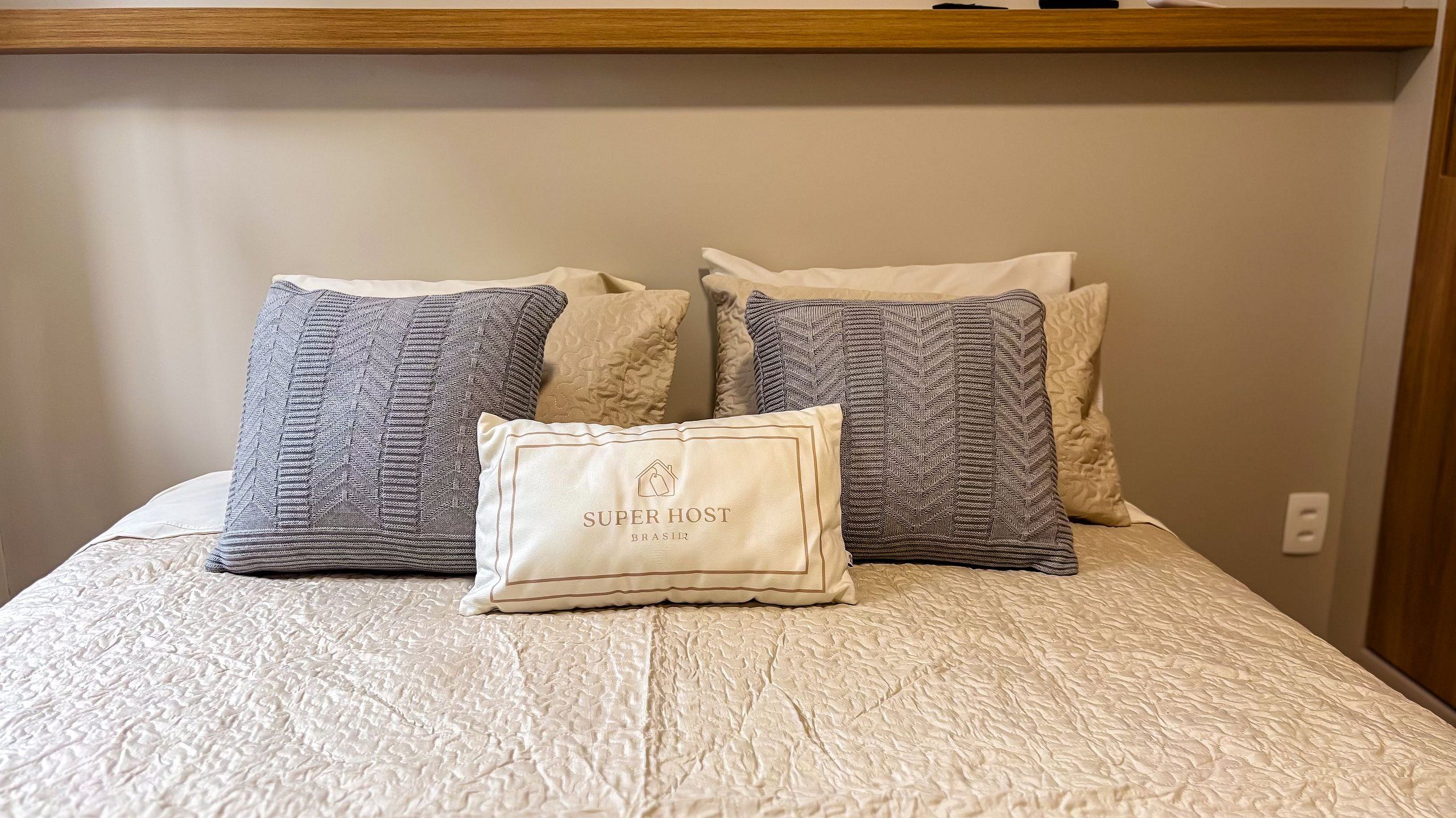 Uma cama com um edredom branco texturizado, duas almofadas estampadas e uma almofada menor com a inscrição 'SUPER HOST BRASIR'.
