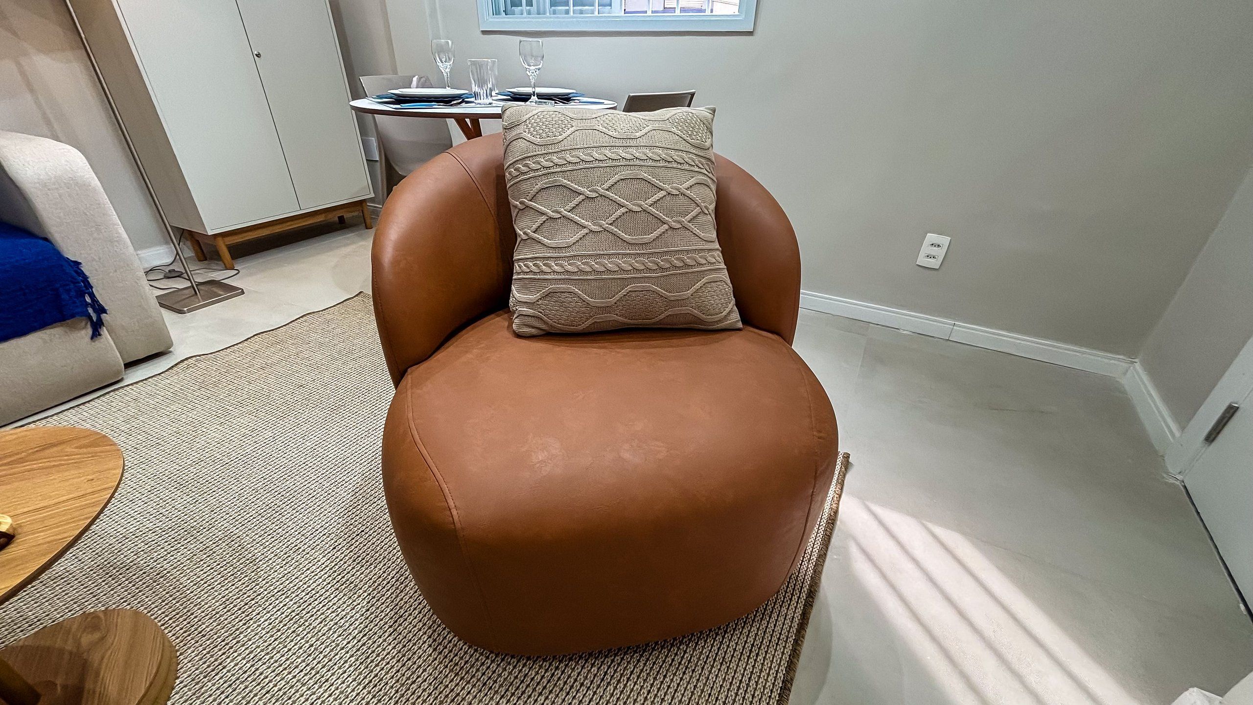 Uma cadeira de couro marrom com uma almofada estampada em um quarto com uma mesa de jantar e uma janela ao fundo.