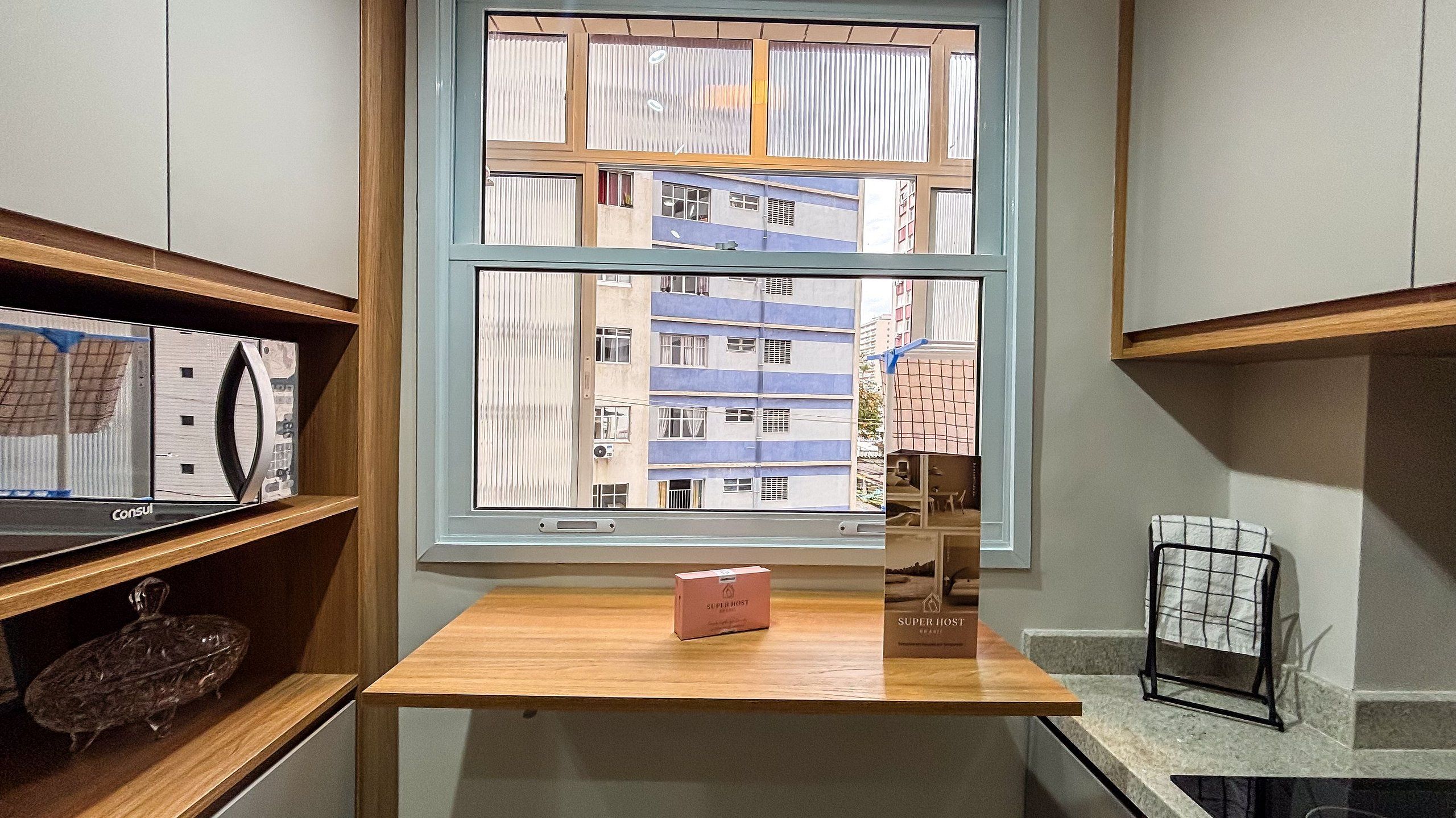 Una habitación con ventana que da a un edificio, una mesa de madera con una caja y estanterías con objetos.
