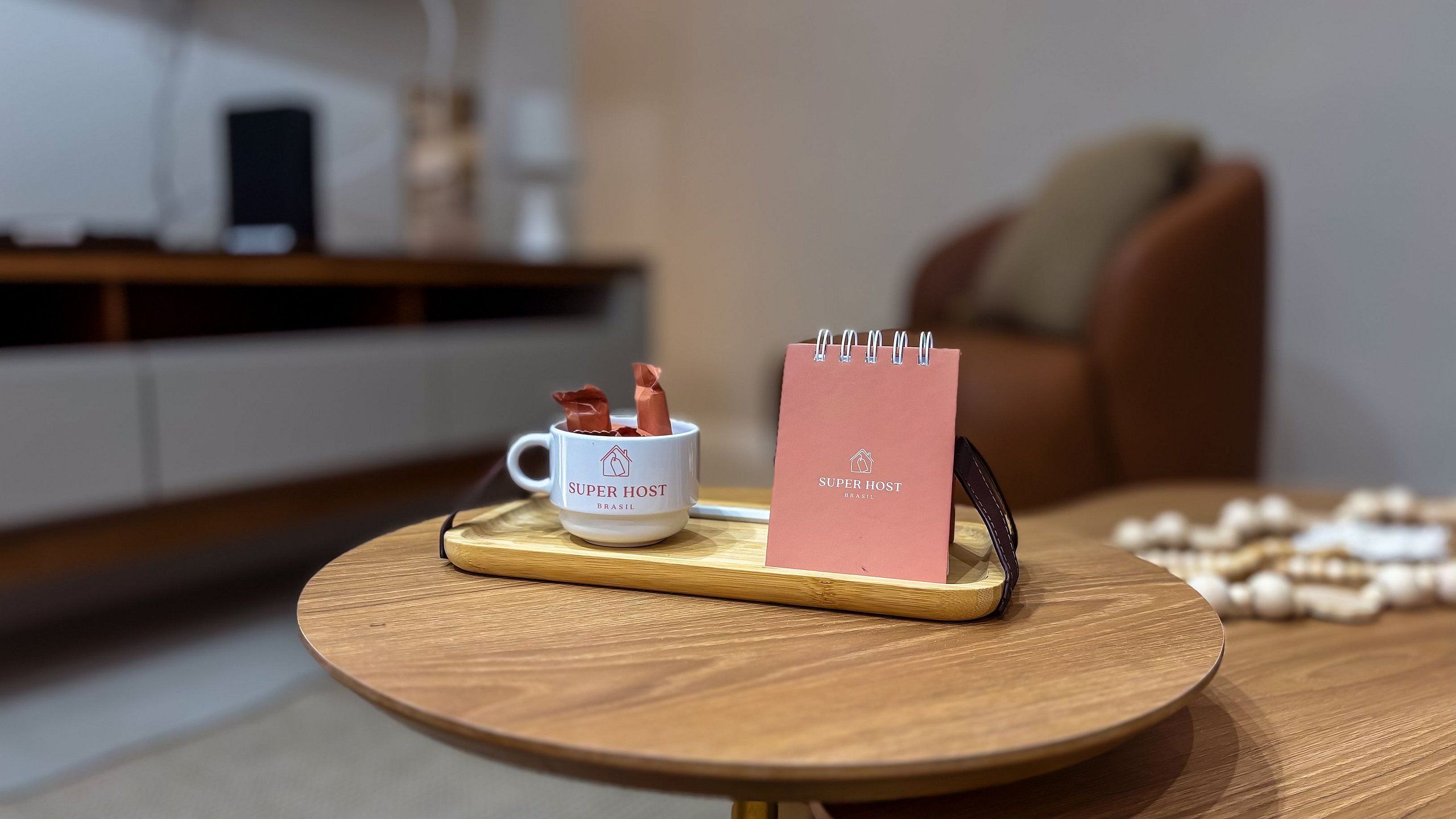 A white mug with 'SUPER HOST BRASIL' on it, a pink notebook with the same branding, and a wooden coaster on a round table.