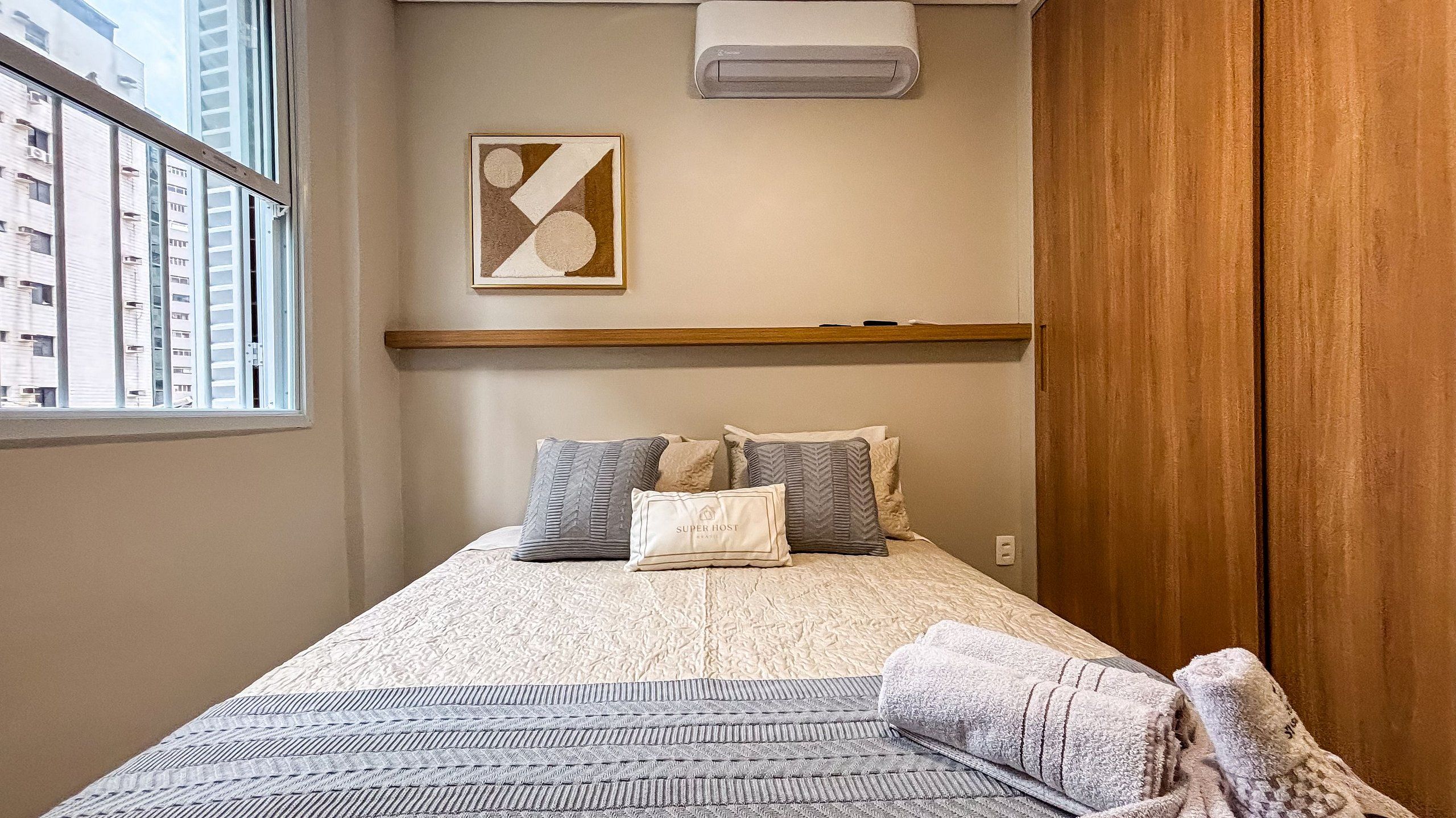 Un dormitorio con cama, almohadas, una colcha, un aire acondicionado de pared y una ventana con vistas a edificios.