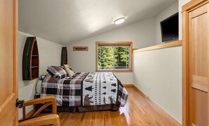 Ein Schlafzimmer mit einem Bett, einem Sessel, einem Fenster mit Blick auf Bäume, einem an der Wand befestigten Fernseher und einem Holzschrank.