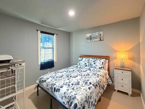 Un dormitorio con cama, una mesita de noche con lámpara, una ventana con persianas y una obra de arte colgada en la pared.