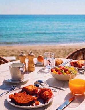 Ett frukostbord dukat upp på en strand med utsikt över havet, med tallrikar fyllda med mat, ett glas apelsinjuice och en skål med frukt.