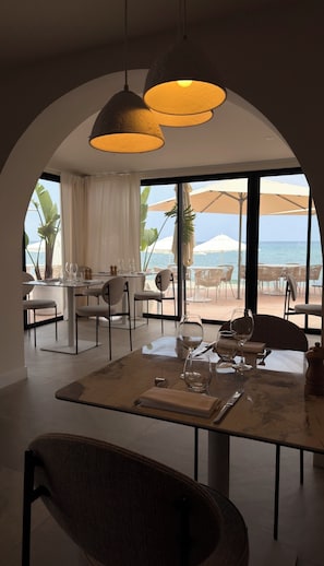 Comedor con una mesa dispuesta para dos personas, con vista a una playa con sombrillas.