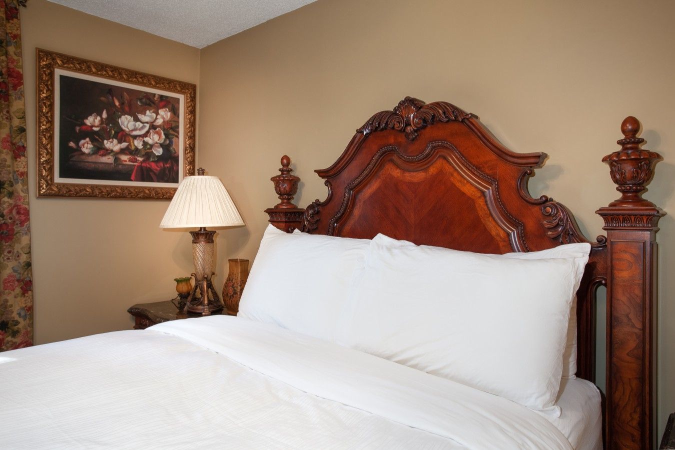 A neatly made bed with a wooden headboard, a bedside lamp, and a framed floral painting on the wall.