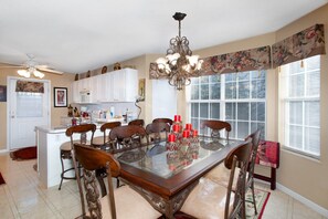 A dining area with a glass table, wooden chairs, a chandelier, and a kitchen in the background. - Moonridge Lodge | Fireplace, Grill & More! (Pigeon Forge)