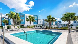 A rectangular swimming pool with a metal ladder, surrounded by lounge chairs and palm trees, with modern buildings in the background.