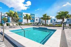 A rectangular swimming pool with a metal ladder, surrounded by lounge chairs and palm trees, with modern buildings in the background.