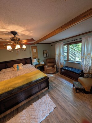 A bedroom with a large bed, a wooden ceiling fan, a window with curtains, a leather chair, and a bench.