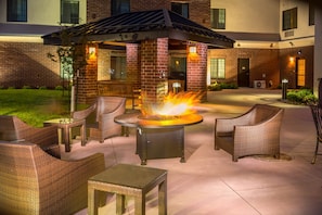 An outdoor patio with wicker furniture, a central fire feature, and a brick building in the background.
