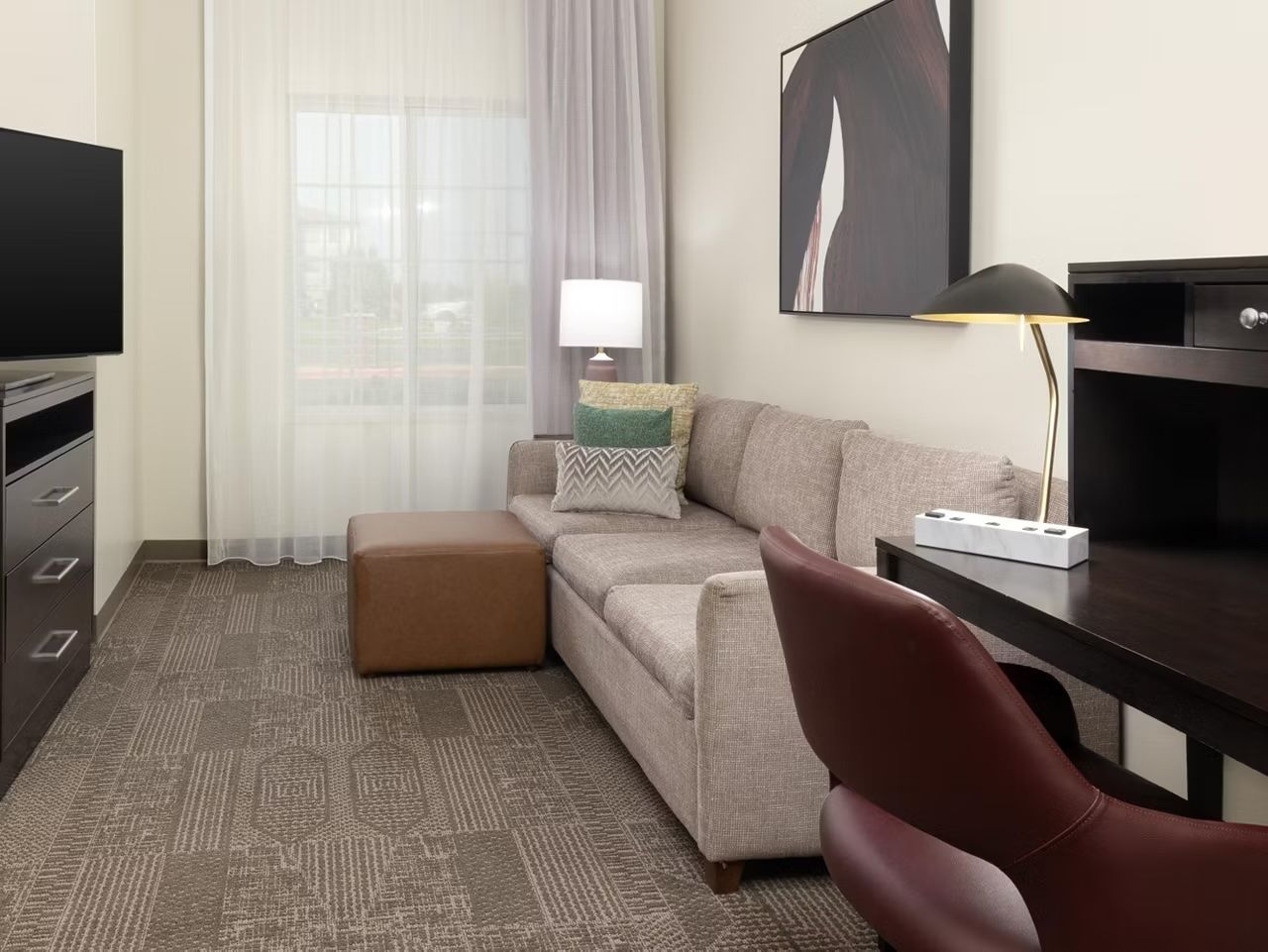 A hotel room with a sofa, ottoman, a desk with a lamp, a TV, and a large window with sheer curtains.