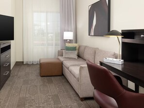 A hotel room with a sofa, ottoman, a desk with a lamp, a TV, and a large window with sheer curtains.