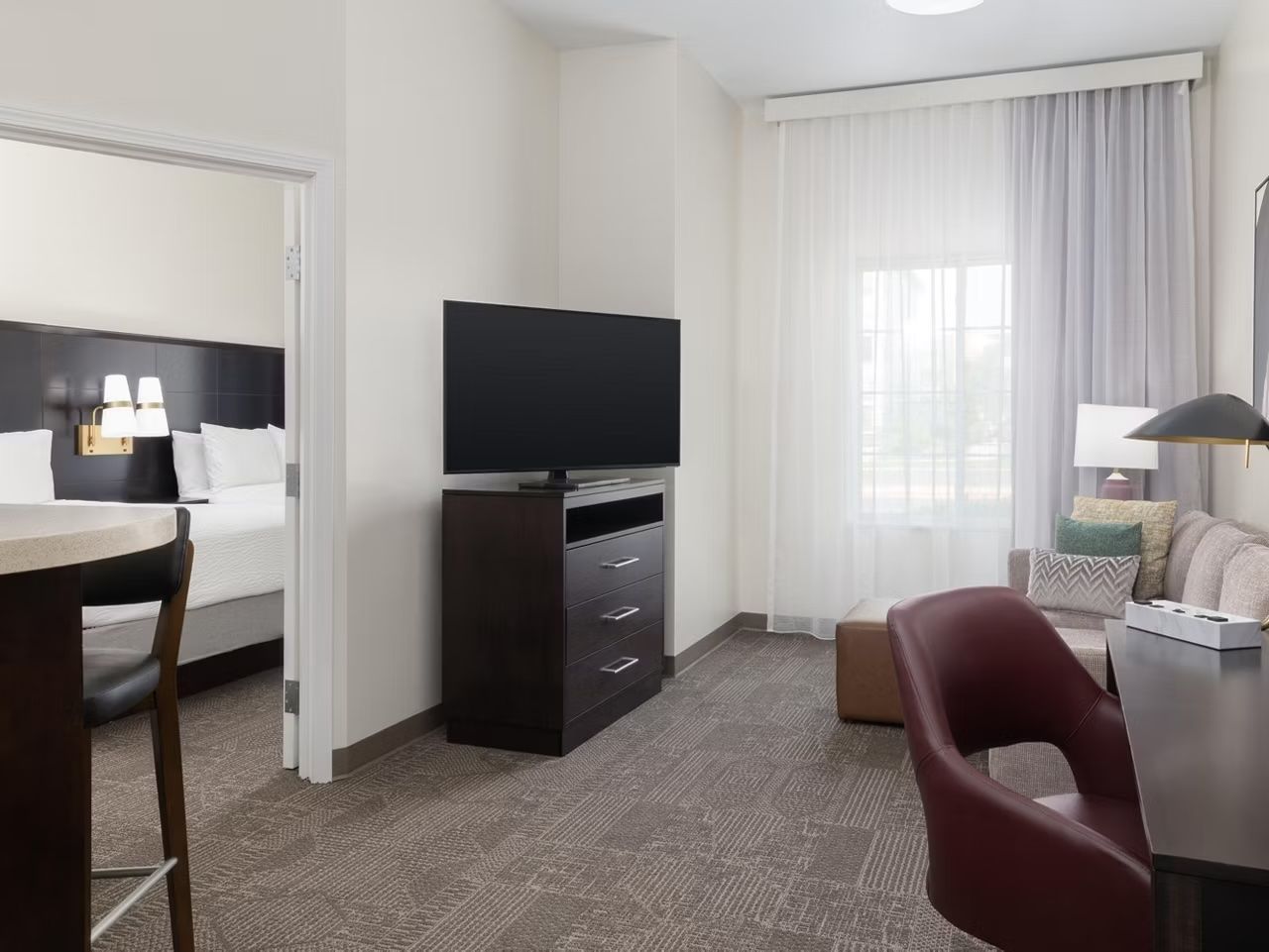 Ein Hotelzimmer mit Bett, Fernseher auf einem Tisch, Schreibtisch mit Stuhl und einem Fenster mit Vorhängen.