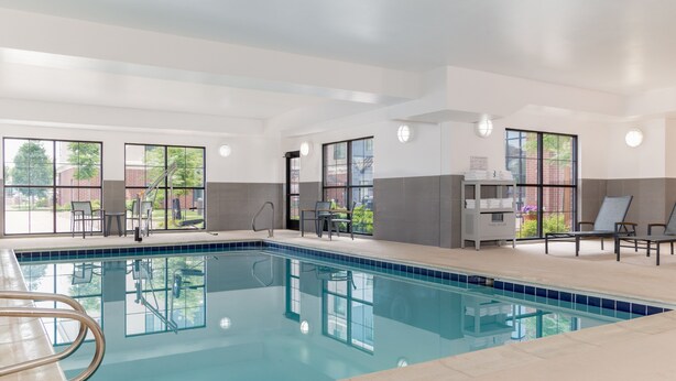 En moderne indendørs swimmingpool med glasvægge, et solbelyst område med siddepladser og en håndklædeafleveringsstation.