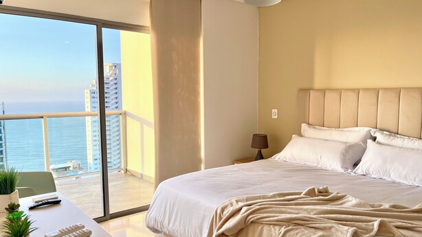 A bedroom with a bed, a desk, a lamp, a potted plant, and a view of the ocean. - by Calamari.Homes - Infinitum 3302 - Bocagrande (Bocagrande)