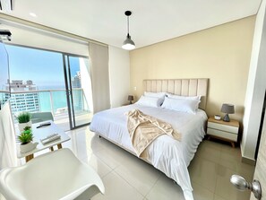 A modern hotel room with a large bed, bedside tables, a desk with a chair, and a view of the city. - by Calamari.Homes - Infinitum 3302 - Bocagrande (Bocagrande)