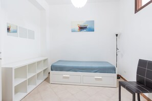 Ein kleines, weißes Zimmer mit einem Einzelbett, einem Sessel, einer Stehlampe und einem Regal.