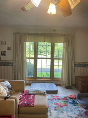 Ein Zimmer mit einer Couch, einem kleinen Hocker, einem bunten Teppich, einer Holzablage und einem Fenster mit Vorhängen.