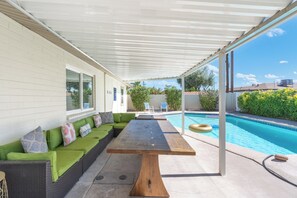 Ein überdachter Außenbereich mit Pool, einem langen Holztisch und einem grünen Sofa mit Kissen.