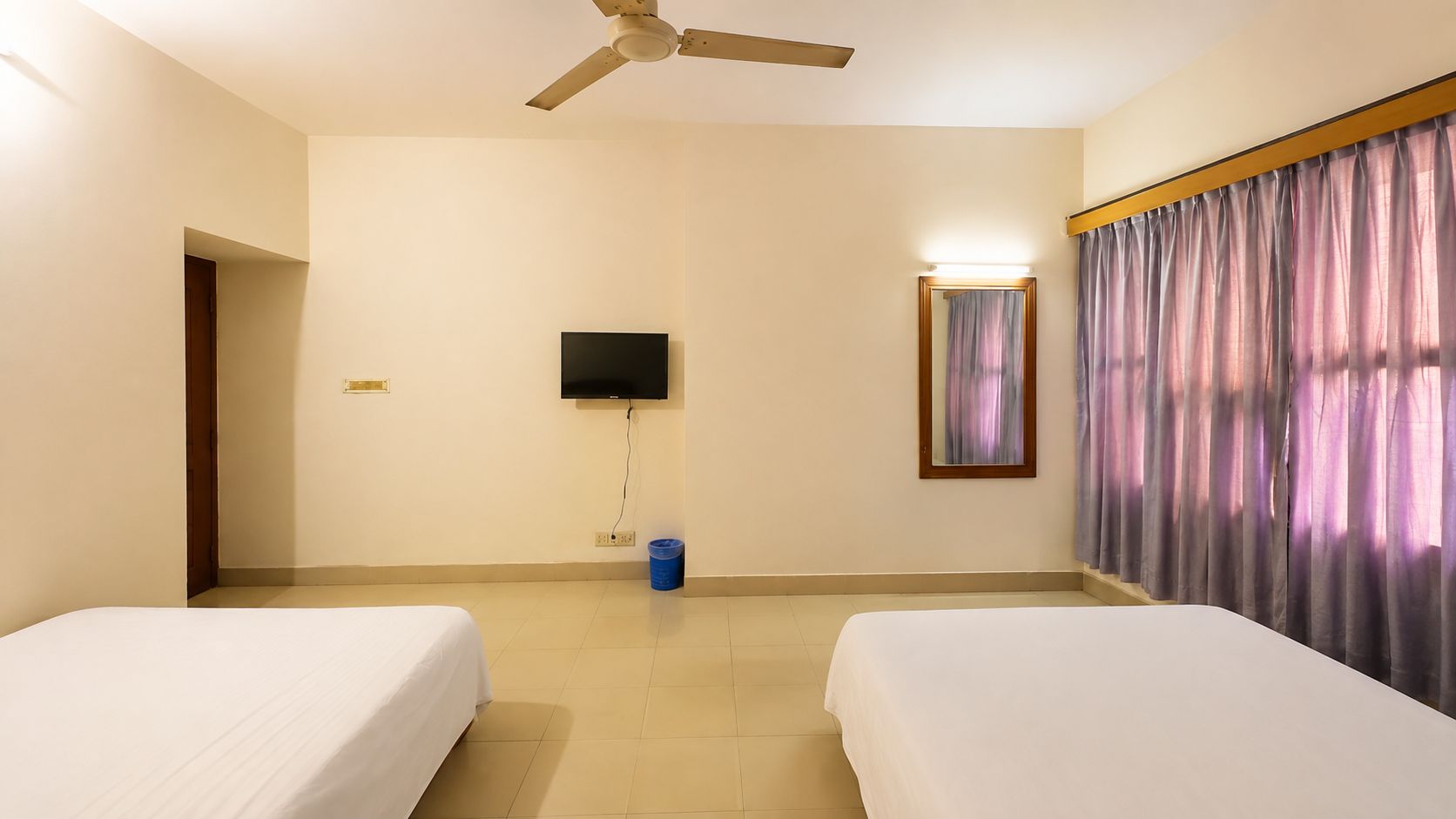 A hotel room with two beds, a ceiling fan, a TV, a mirror, and a curtain.