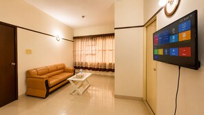 Una sala de estar con un sofá de cuero marrón, una mesa de centro blanca, un televisor de pantalla plana montado en la pared mostrando varios canales, y una ventana con cortinas translúcidas.