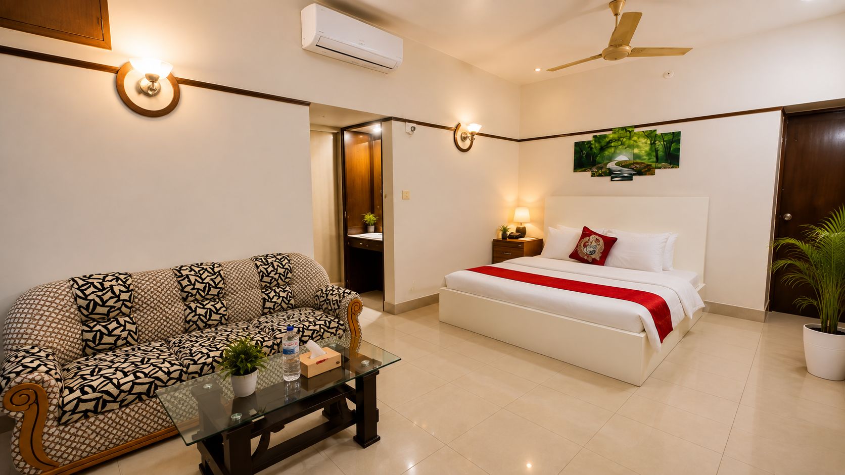 A hotel room with a sofa, a bed with a red blanket, a bedside table with a lamp, and a ceiling fan.